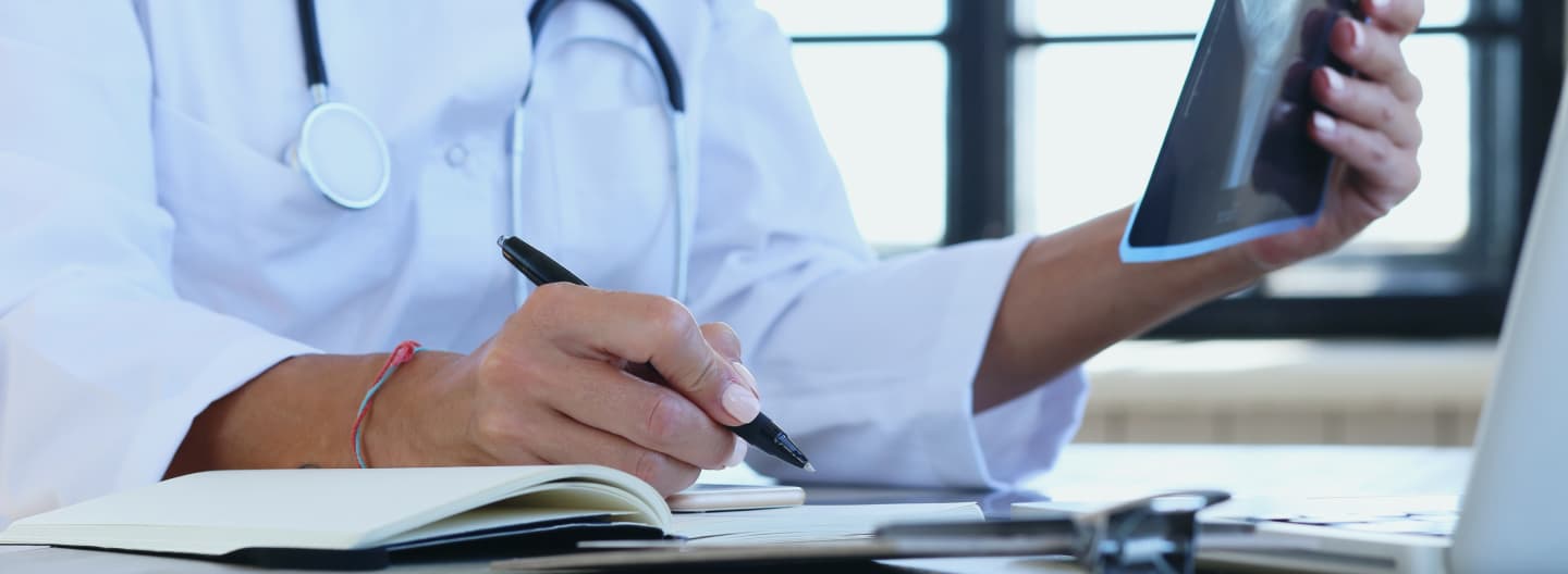  An archaeologist wearing a lab coat is writing in a notebook while holding an x-ray of a bone up to the light.